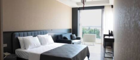 A modern hotel room with a large bed, a seating area with a sofa and armchairs, a small table, and a balcony with a view.