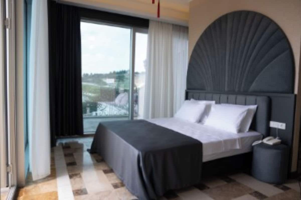 Quarto de hotel moderno com uma cama grande, vista para o exterior e um detalhe arquitetônico único acima da cama.