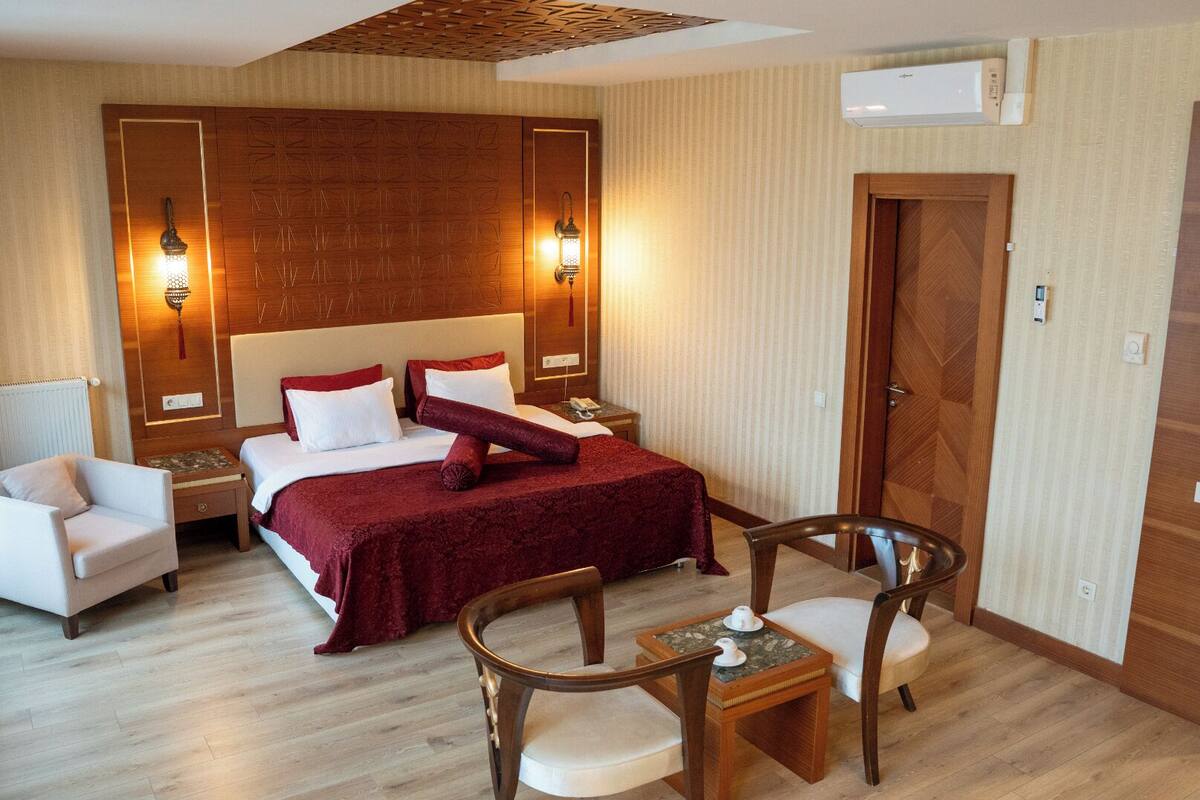 Quarto de hotel com uma cama grande, duas mesas de cabeceira, um painel de cabeceira de madeira, uma porta de madeira e uma pequena mesa com cadeiras.