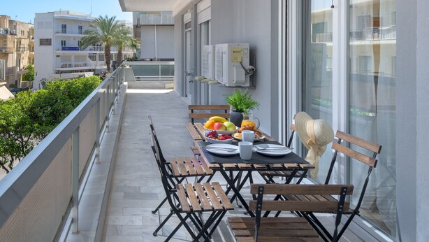 A balcony with a table set for breakfast, including fruits, pastries, and a beverage. There are chairs and a sunshade.
