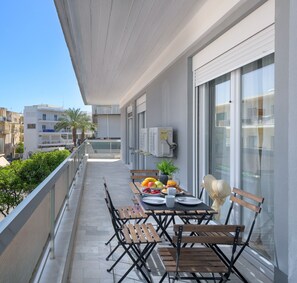 A balcony with a table set for breakfast, including fruits, pastries, and a beverage. There are chairs and a sunshade.