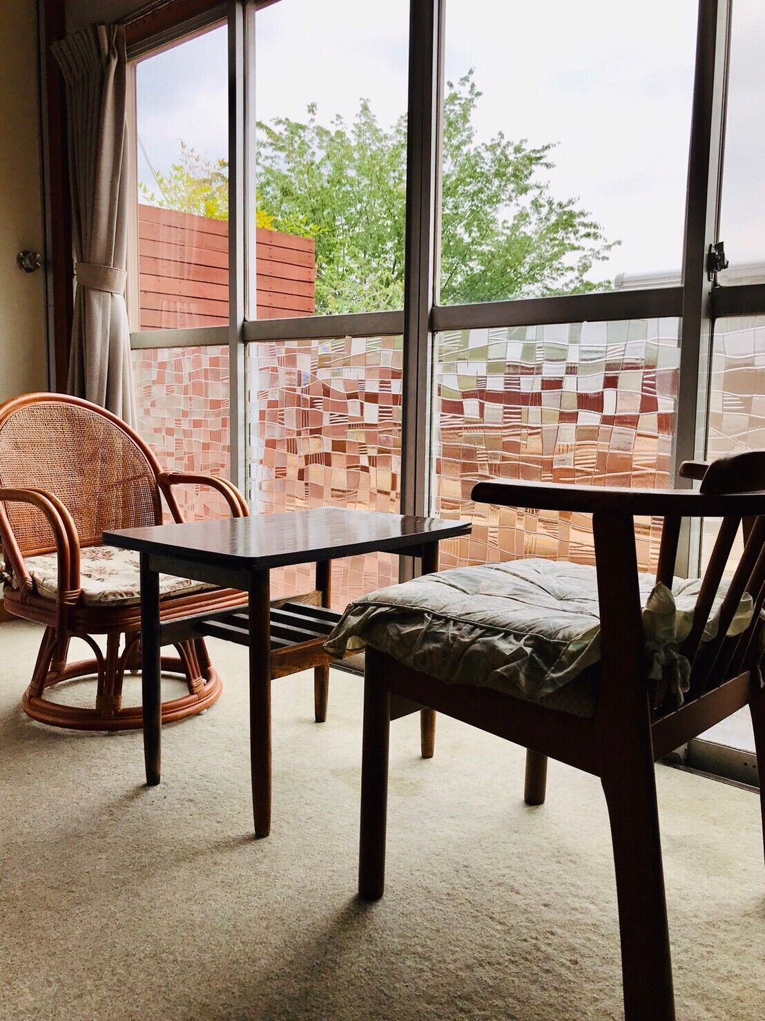 Ein Zimmer mit großem Fenster, Korbstühle, einem Holztisch und Blick auf eine Ziegelwand und Grünflächen.