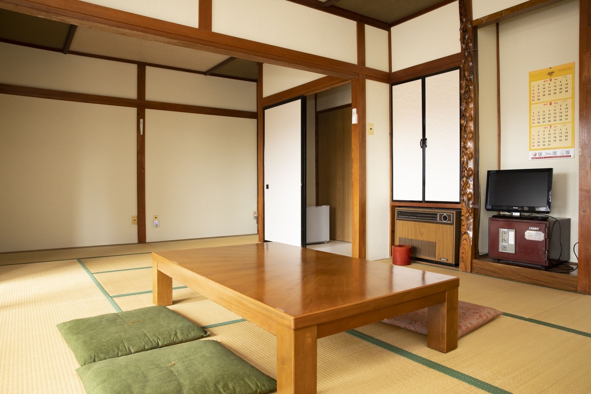 Een traditionele Japanse kamer met tatami vloeren, een houten tafel, groene kussens, een televisie en een agenda aan de muur.