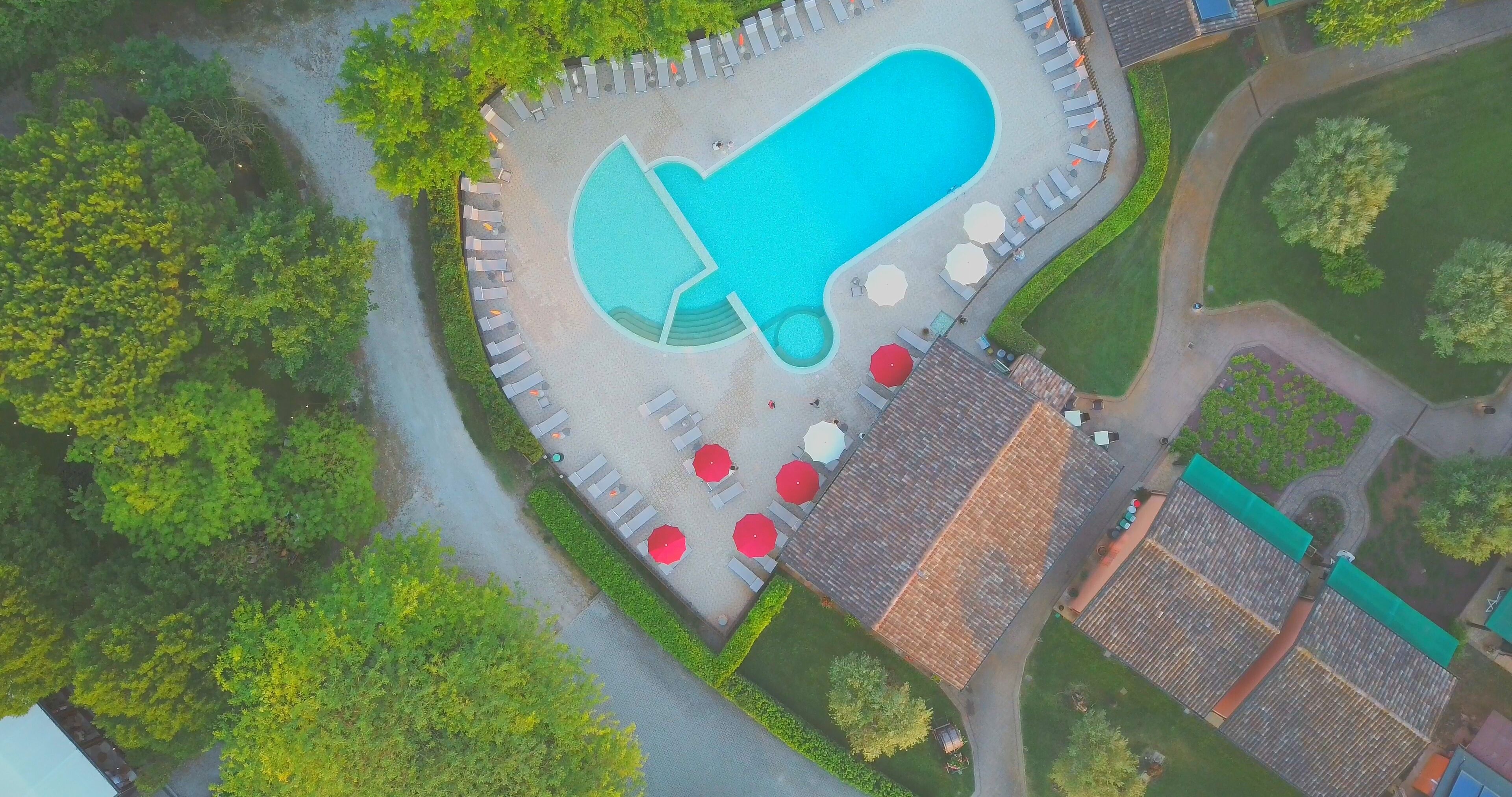 Une vue aérienne d’une piscine à la forme originale, entourée de chaises longues et de parasols, avec des bâtiments adjacents et de la végétation.