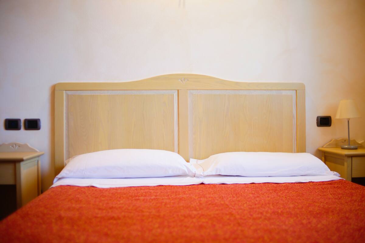 A hotel room with a wooden headboard, a red bedspread, and a nightstand with a lamp.