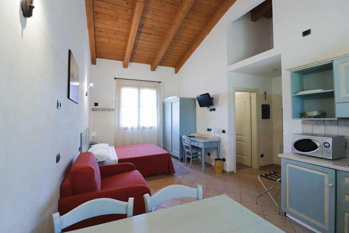A compact room with a wooden ceiling, a dining table, two red beds, a desk, a television, and a kitchenette.