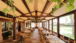 Un restaurante con mesas y sillas de madera, ventanas amplias y plantas colgantes.