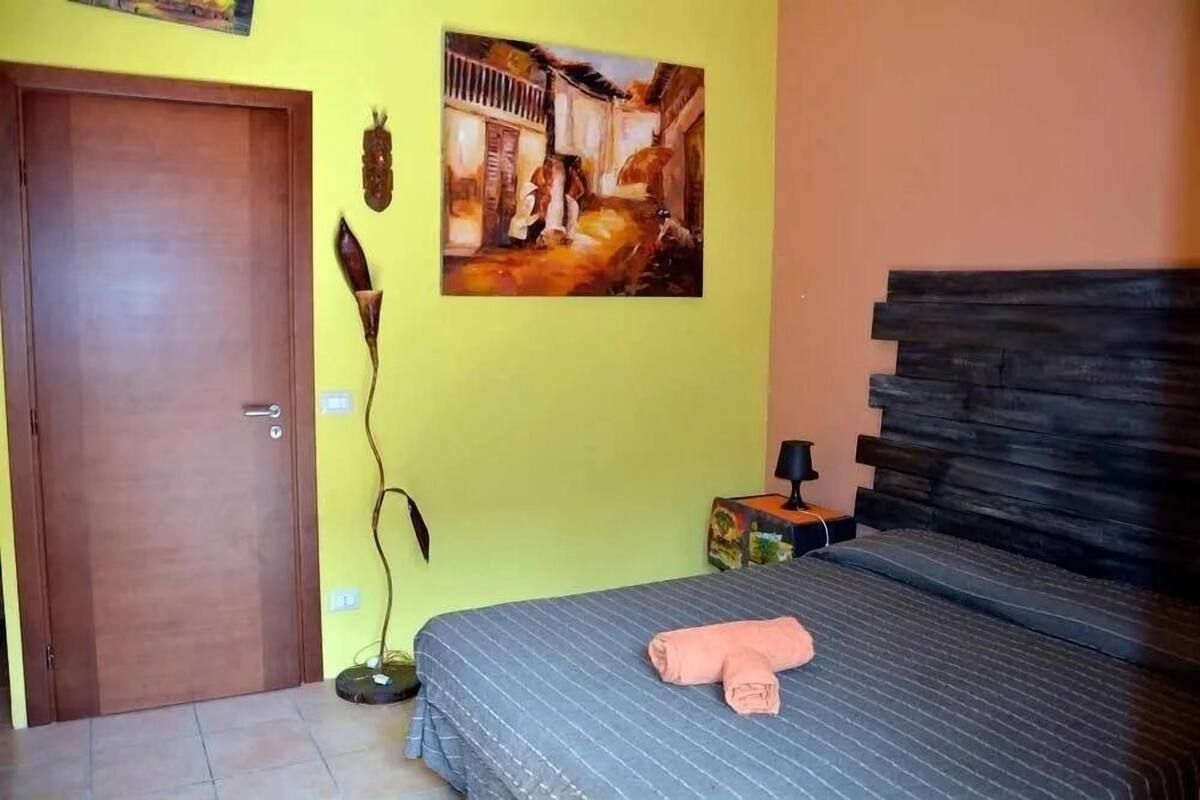 Una habitación con cama, puerta, un cuadro y una mesita de noche.