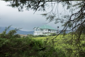 A large, multi-story house with a green roof, surrounded by lush greenery and hills in the background. - Grand Camellia (Nuwara Eliya)