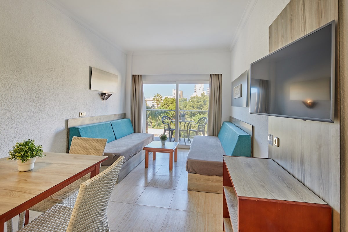 Habitació d'hotel moderna amb sofà, taula de menjador, televisió i un balcó amb vista a edificis i arbres.
