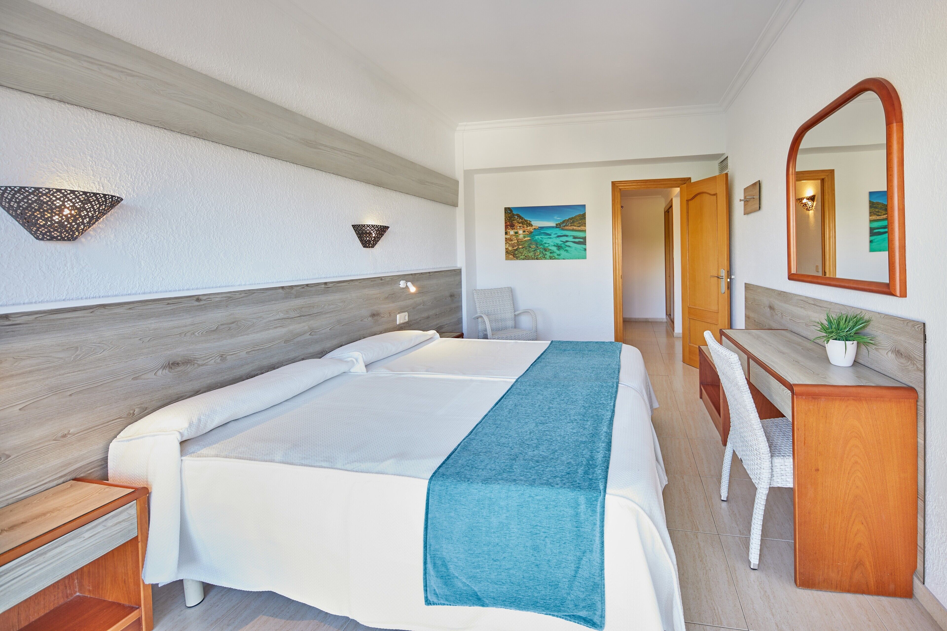 A hotel room with a bed, a desk, a chair, a mirror, and a wall-mounted lamp.