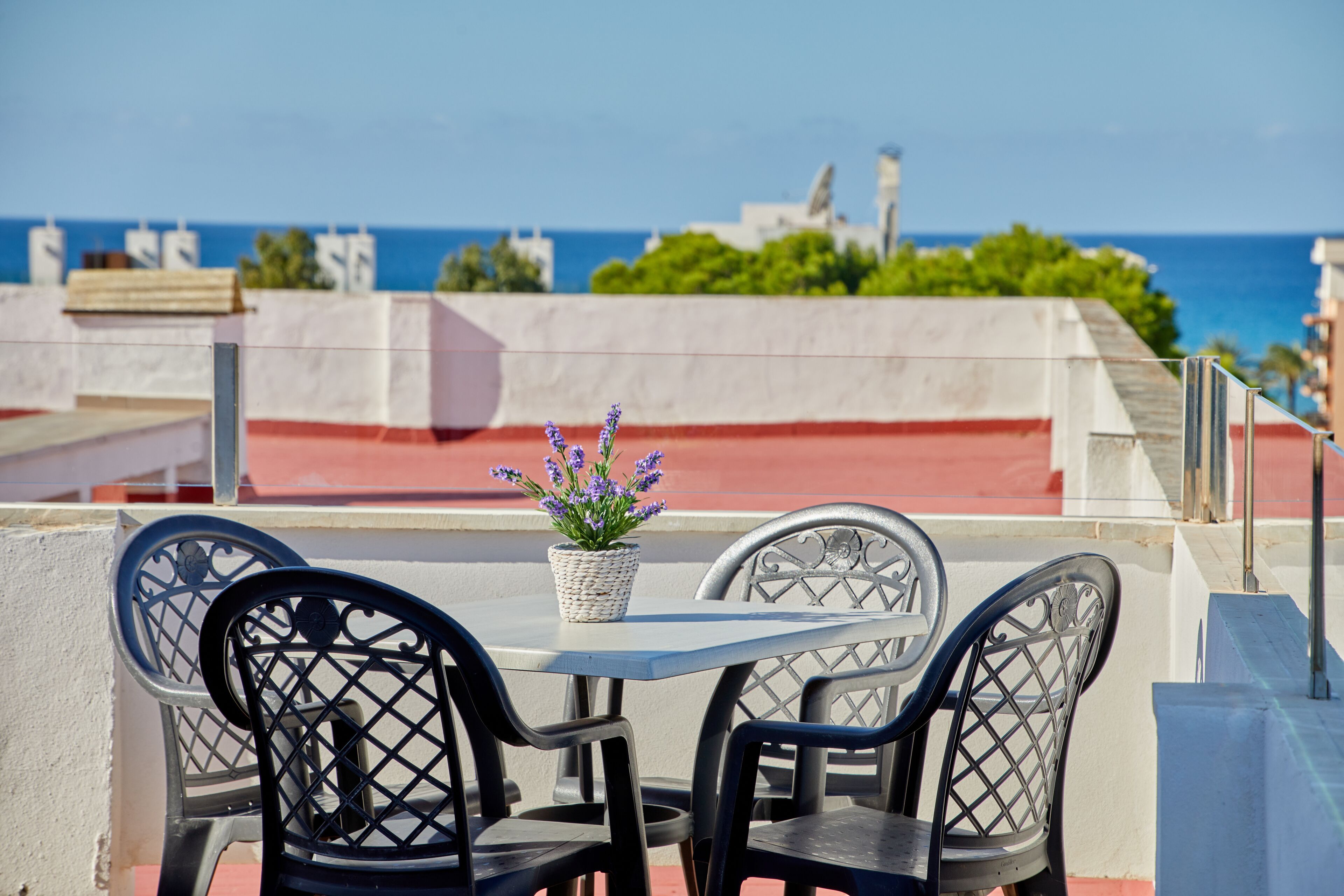 An outdoor dining area with a table and chairs, a potted plant, and a view of the ocean and buildings in the background.