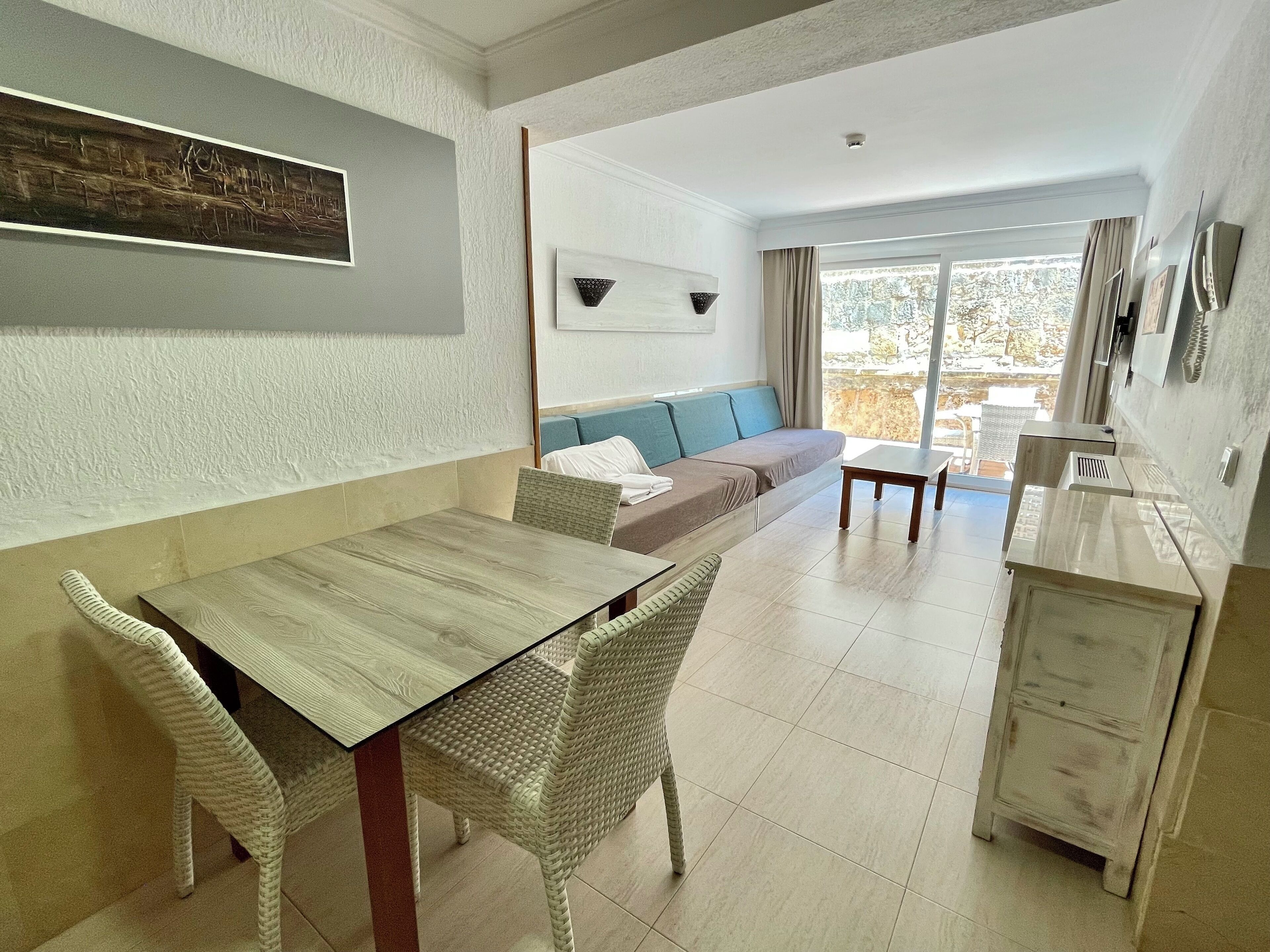 Un área de comedor con una mesa de madera y sillas de mimbre, un sofá con cojines y vistas al exterior a través de un amplio ventanal.
