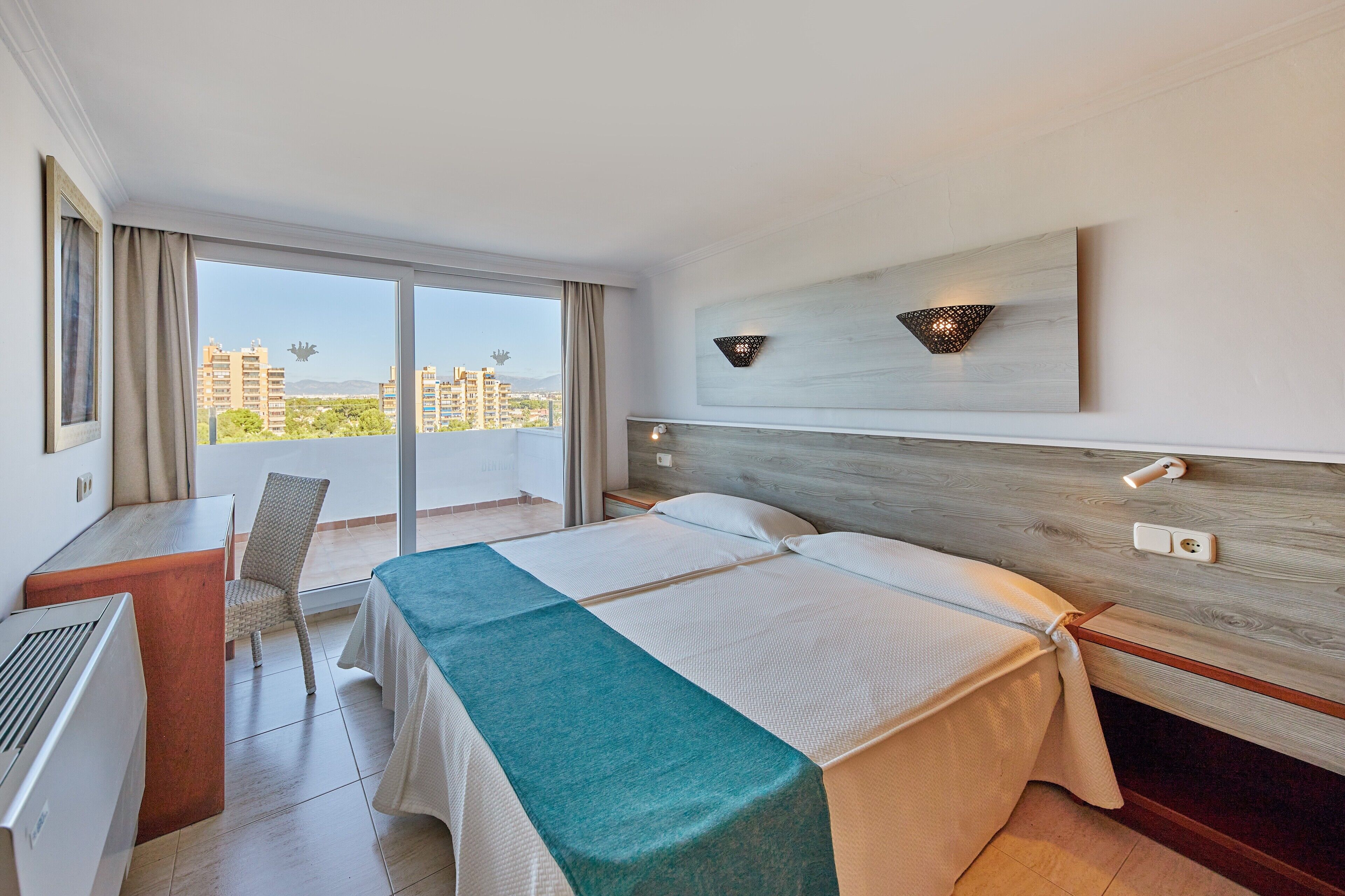 Une chambre d’hôtel comprenant un lit, un bureau, une chaise, une grande fenêtre donnant sur des immeubles et un luminaire fixé au mur.
