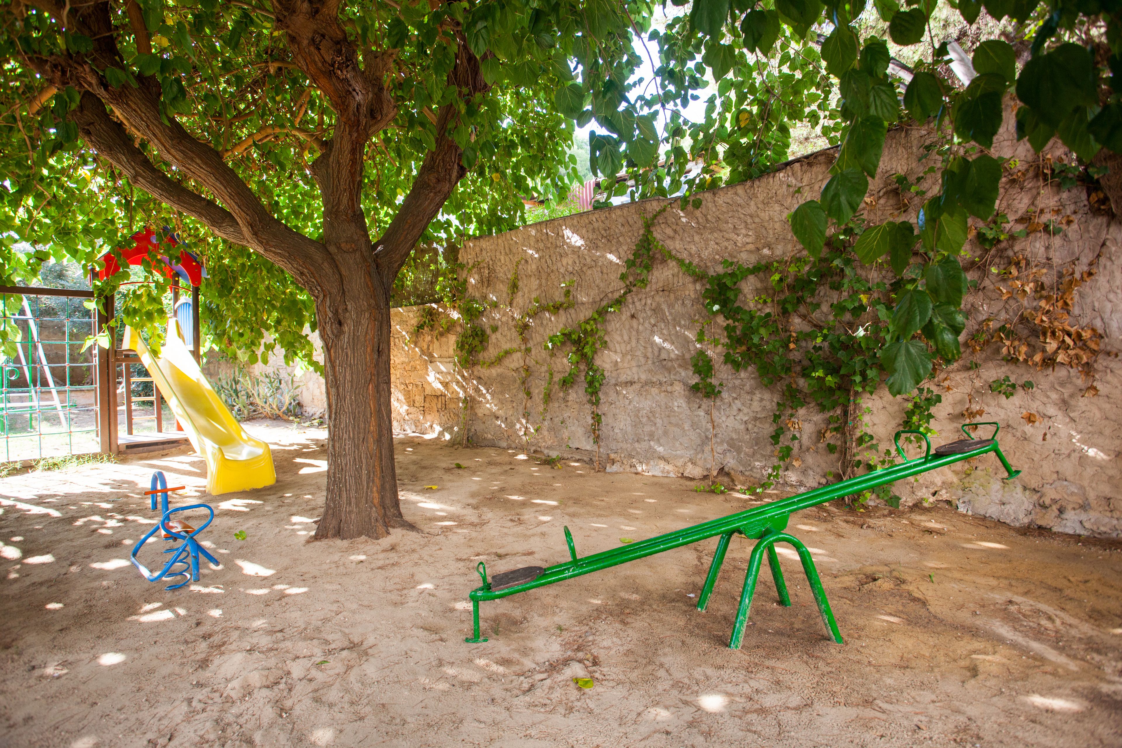Un parque infantil con un sube y baja, un tobogán y un árbol que da sombra.