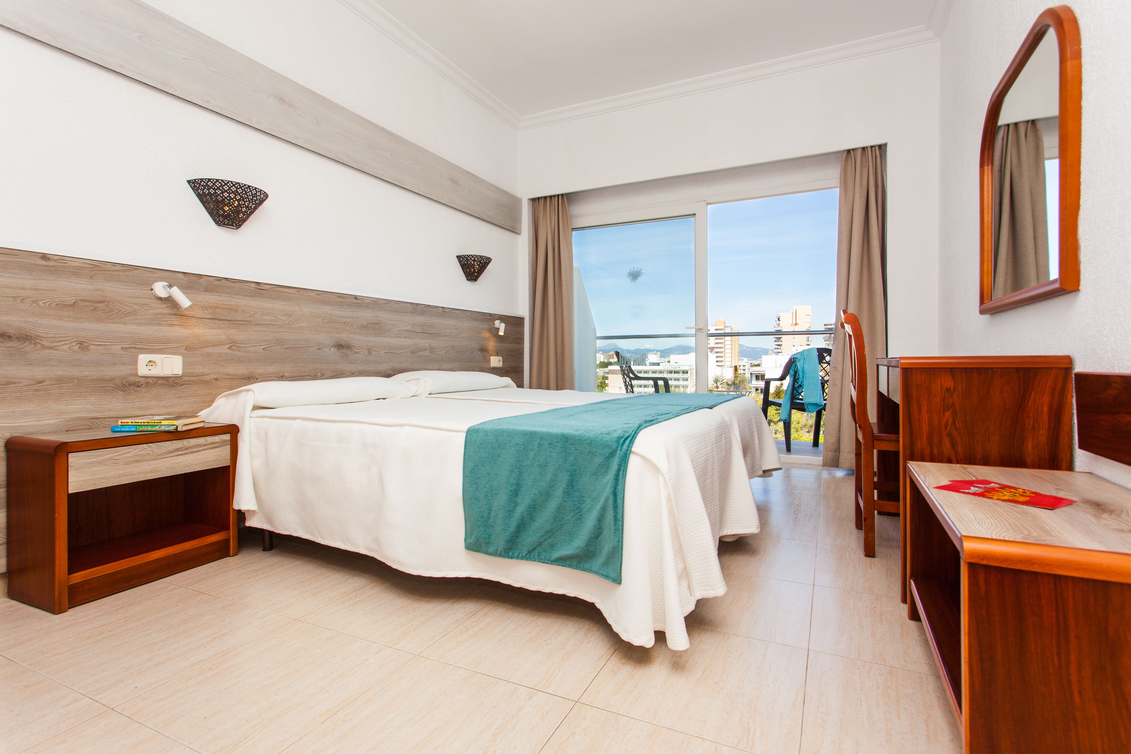 A hotel room with a bed, a desk, a chair, a nightstand, a window with a view, and a mirror on the wall.