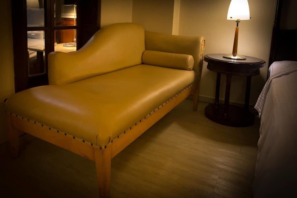 A single bed with a wooden frame and a headboard, a bedside table with a lamp, and a window with blinds.