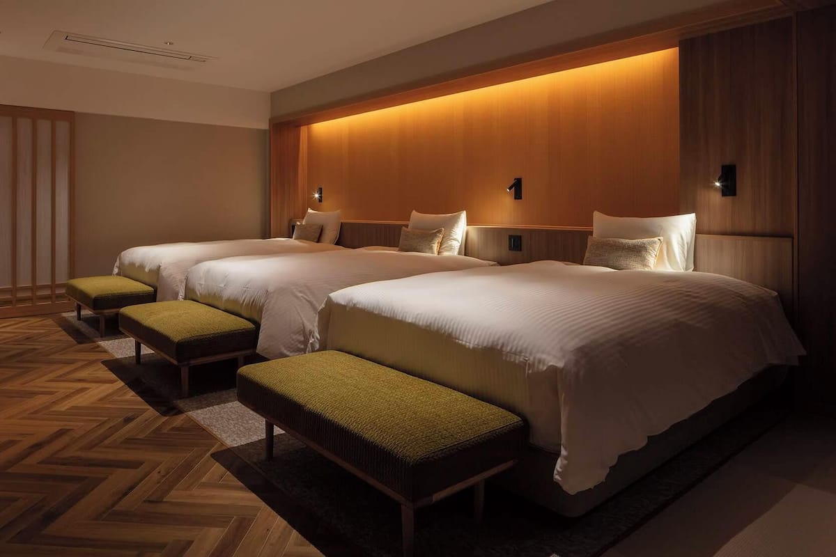 A hotel room with three beds, each with a matching bench. The room has a wooden floor and a warm, illuminated wall.