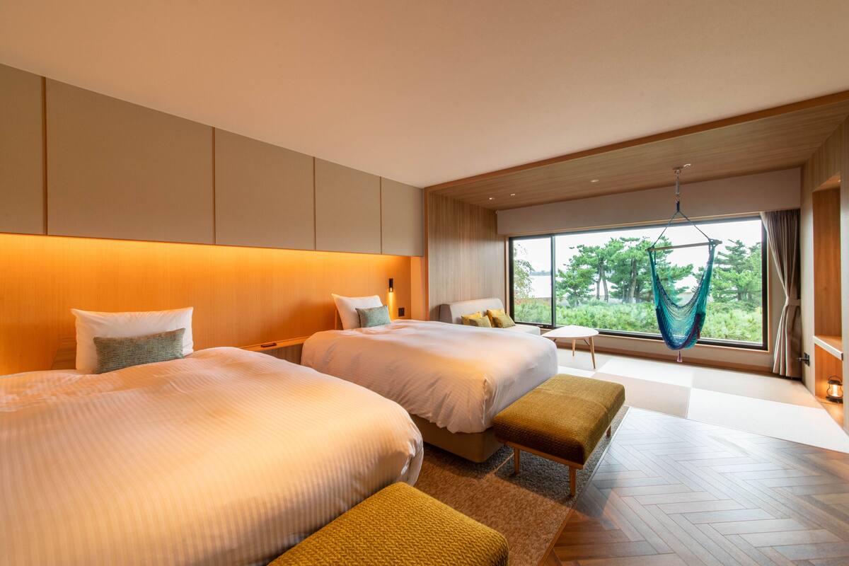 A modern hotel room with two large beds, a hammock, and a view of greenery through the window.