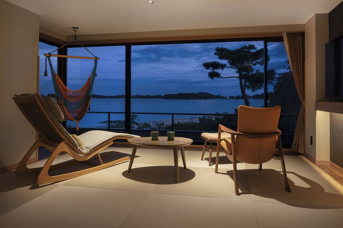 A modern room with a large window, a hammock, two chairs, a small table, and a TV.