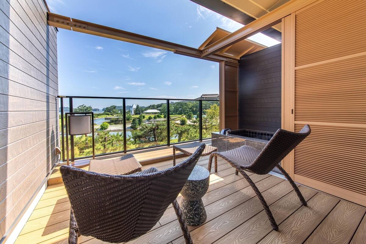 A balcony with wicker chairs, a table, and a view of a river and greenery.