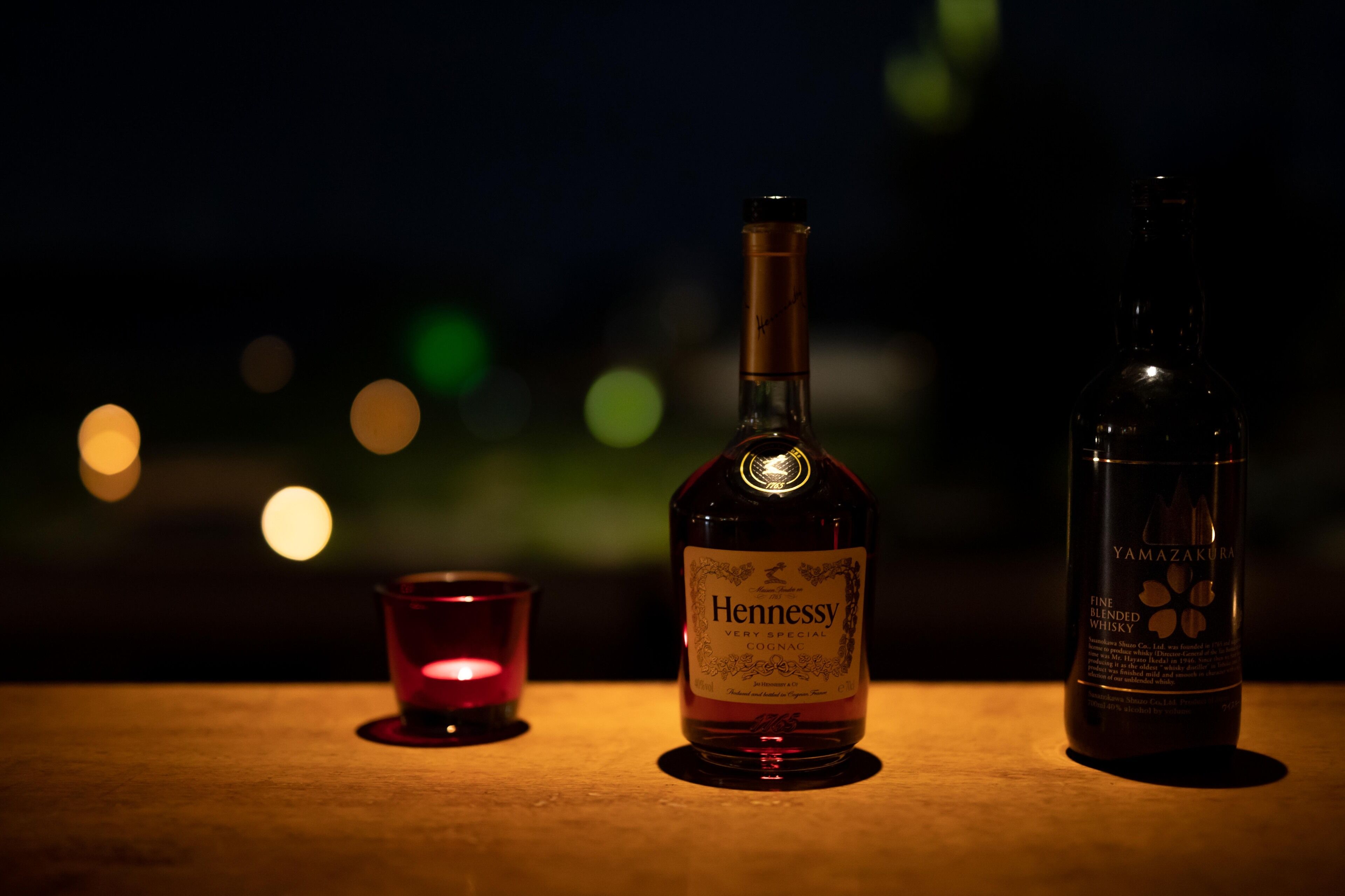 A bottle of Hennessy Very Special Cognac and a bottle of Yamazakura Fine Blended Whisky on a wooden surface with a blurred background of lights.