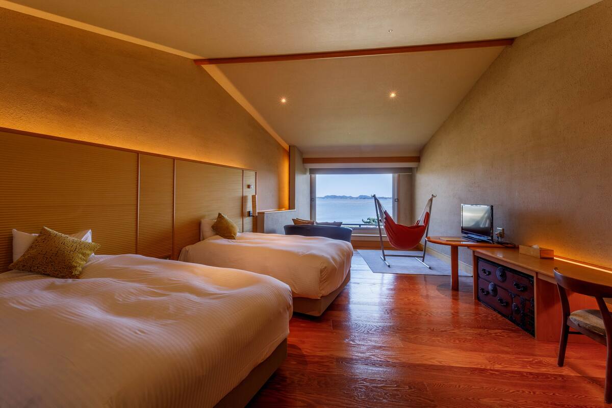 A hotel room with two beds, a television, a desk, and a view of the sea.