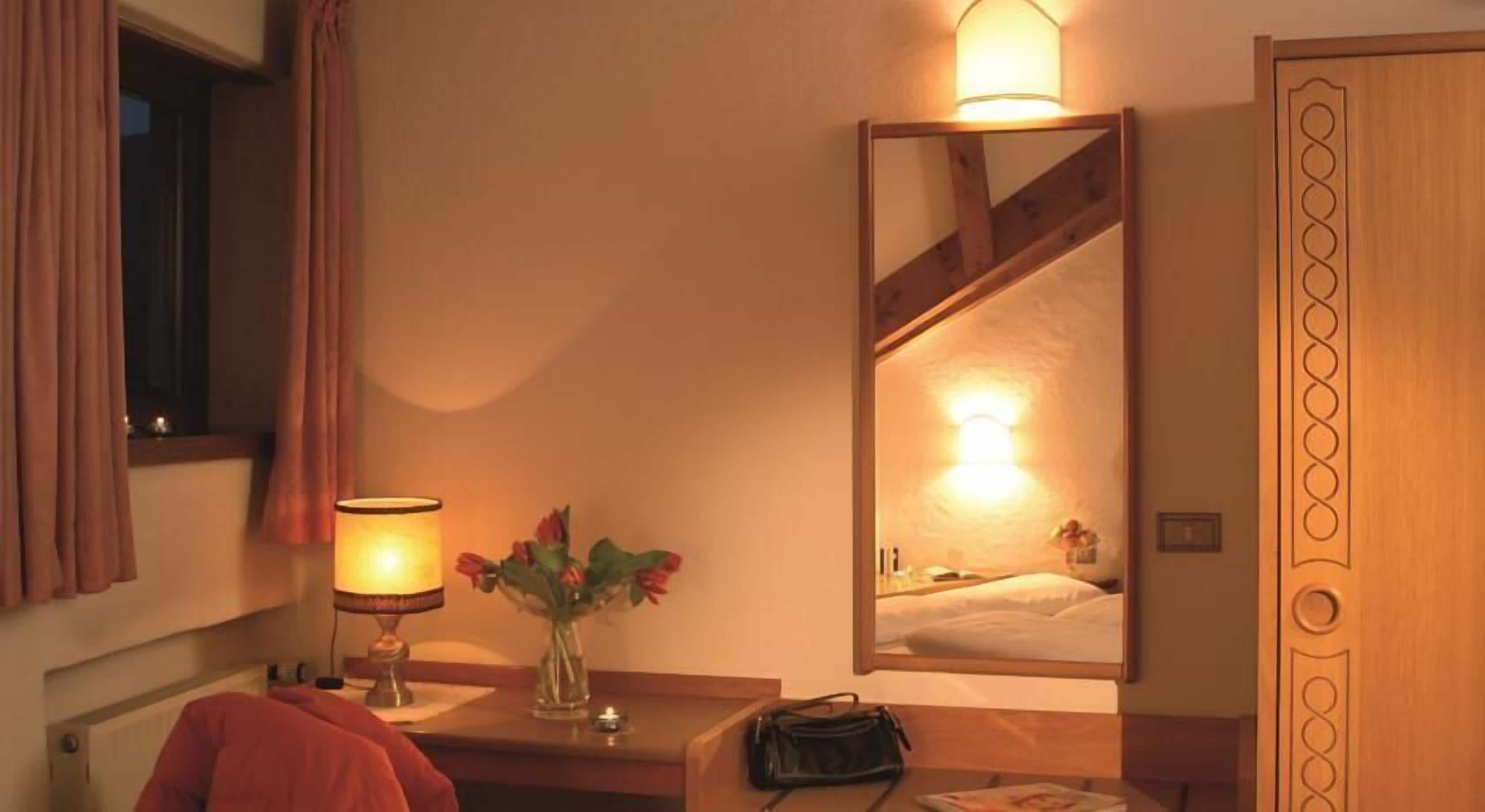 A hotel room with a bed, a bedside table with a lamp, a vase with flowers, and a mirror reflecting the room.