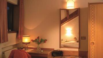 A hotel room with a bed, a bedside table with a lamp, a vase with flowers, and a mirror reflecting the room.