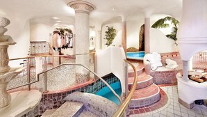 Área de piscina interior luxuosa, com uma banheira de hidromassagem central, vários níveis de escadas e elementos arquitetônicos clássicos como colunas e arcos.