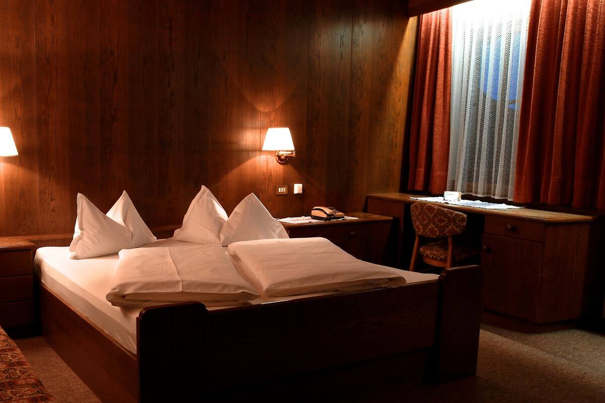 Habitación de hotel con piso de madera, cama con ropa blanca, dos lámparas de noche, mesita de noche, una silla y ventana con cortinas.
