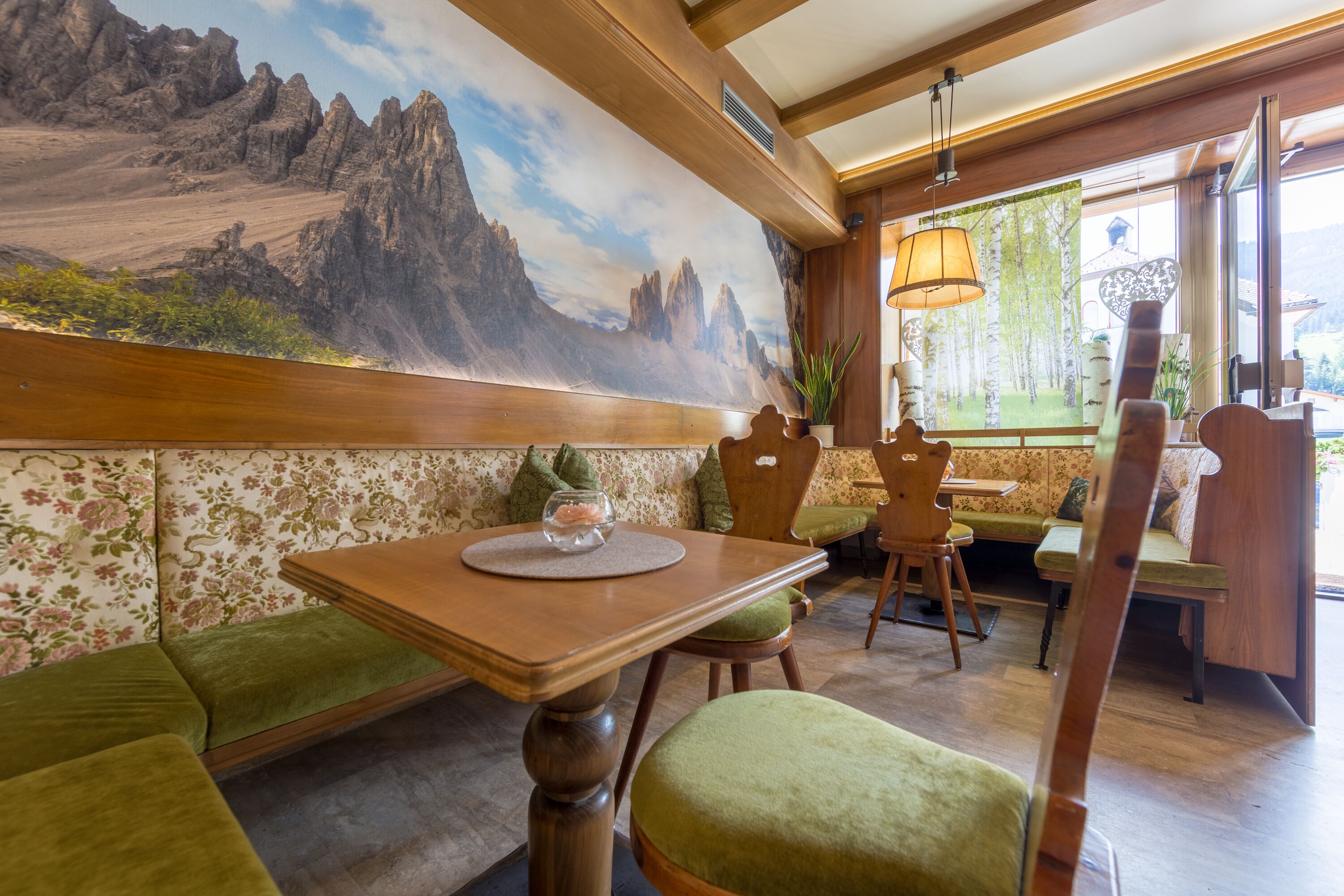 A restaurant interior with wooden furniture, a mural of mountains, and a view of greenery outside.