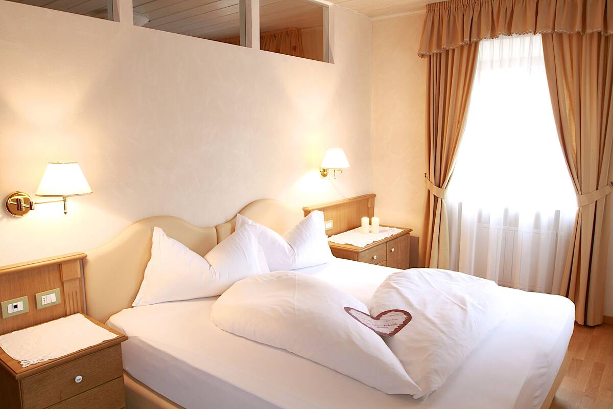 Una cama bien tendida con sábanas y almohadas blancas, dos lámparas de noche, una mesita de madera y una ventana con cortinas.