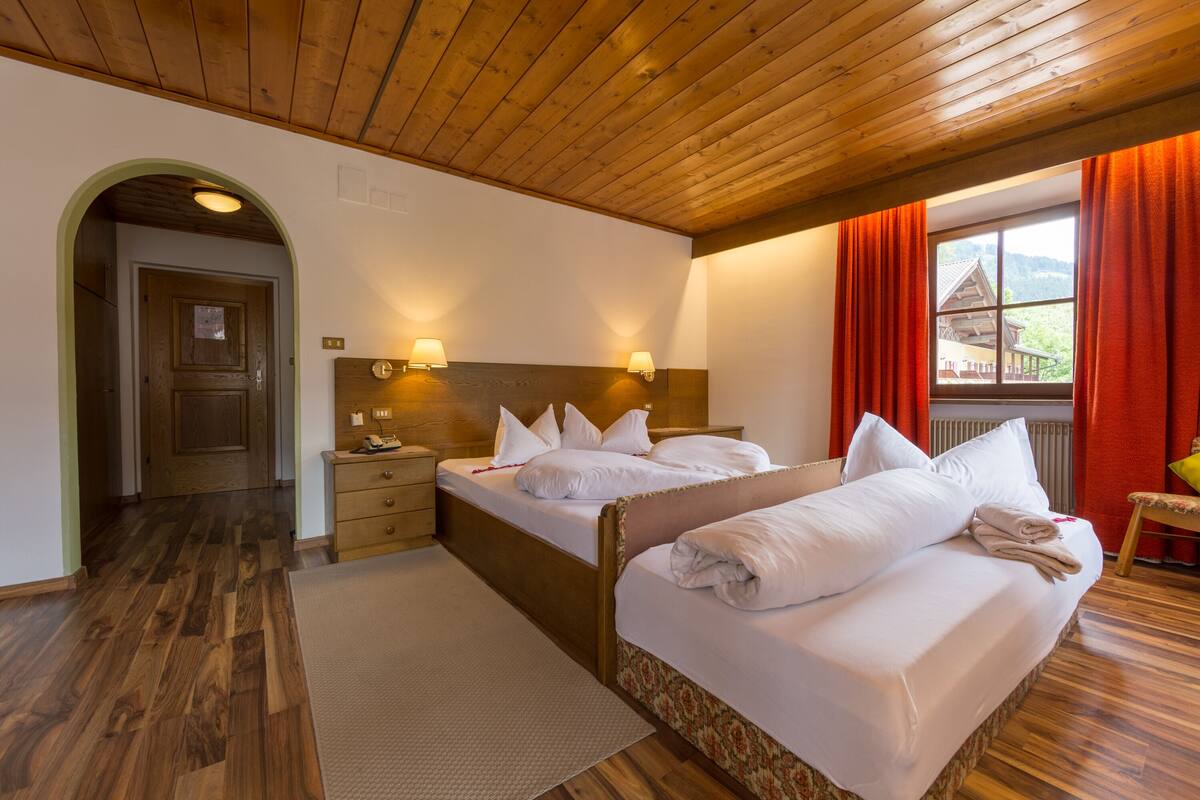 Habitación de hotel con dos camas, piso y techo de madera, una ventana con cortinas rojas y vista a un edificio.