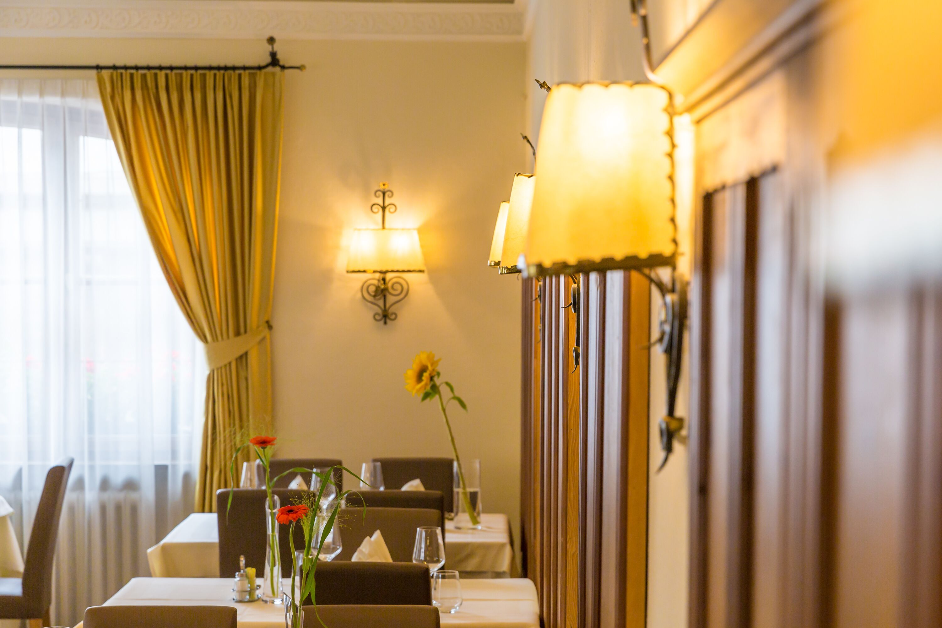 Un coin repas avec des tables préparées pour deux personnes, une lampe fixée au mur et une fenêtre avec des rideaux.