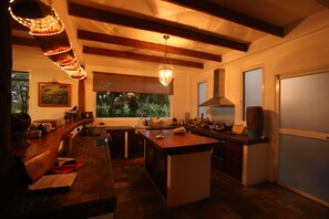 A modern kitchen with a central island, wooden beams, a hanging light fixture, and a large window providing a view of greenery outside. - Semilla Verde Boutique Hotel (Bella Vista)