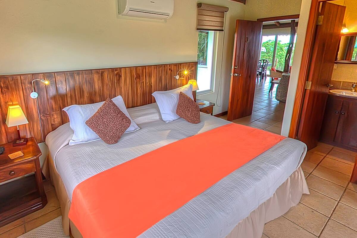 Une chambre à coucher avec une tête de lit en bois, deux lampes de chevet, un lit recouvert d’une couvre-lit blanche et orange, et une vue sur un balcon.