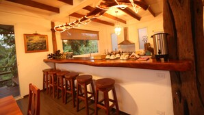 Une cuisine avec des tabourets de bar en bois, un luminaire suspendu et une vue sur l’extérieur.