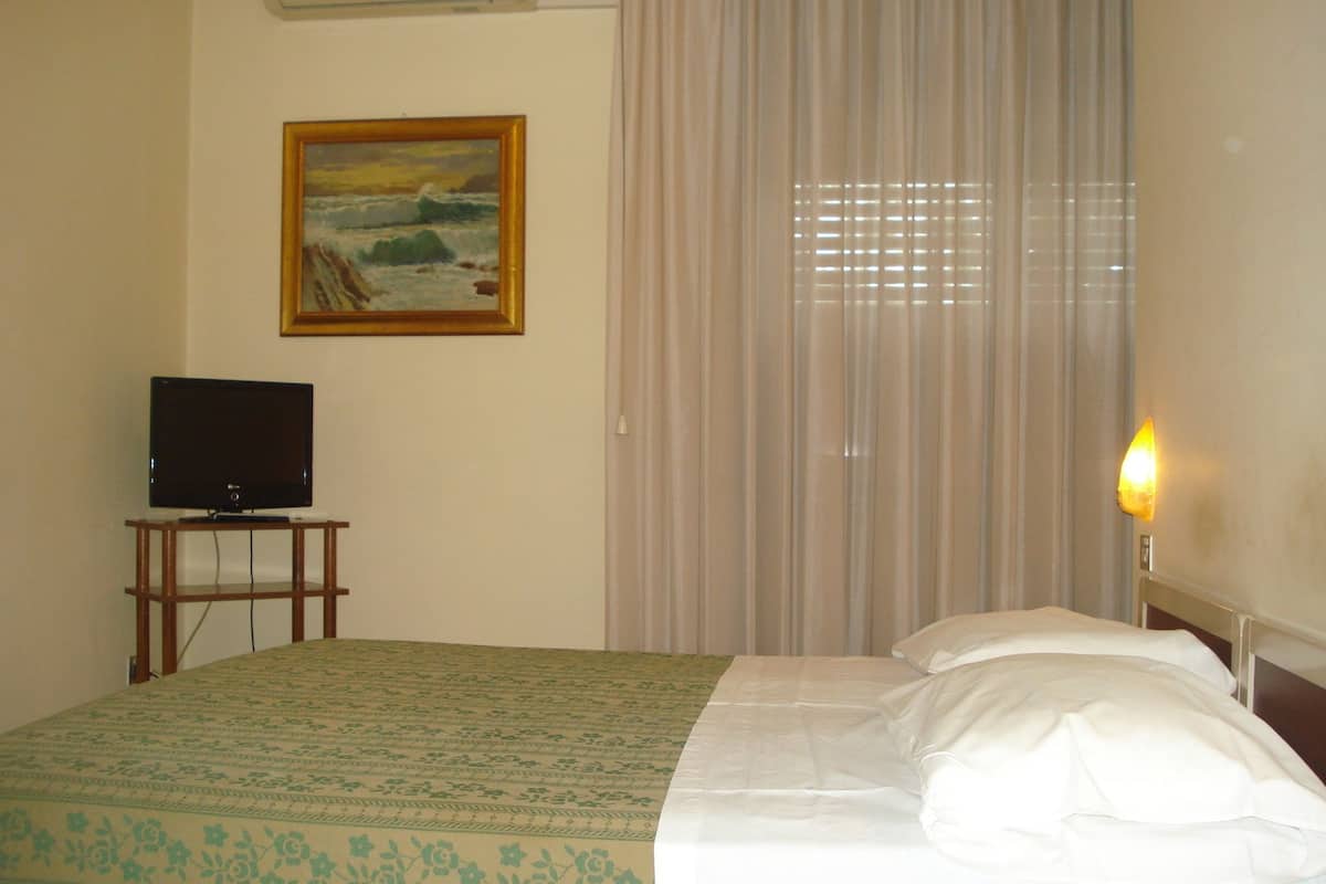 A hotel room with a bed, a television on a wooden stand, a painting on the wall, and a window with curtains.