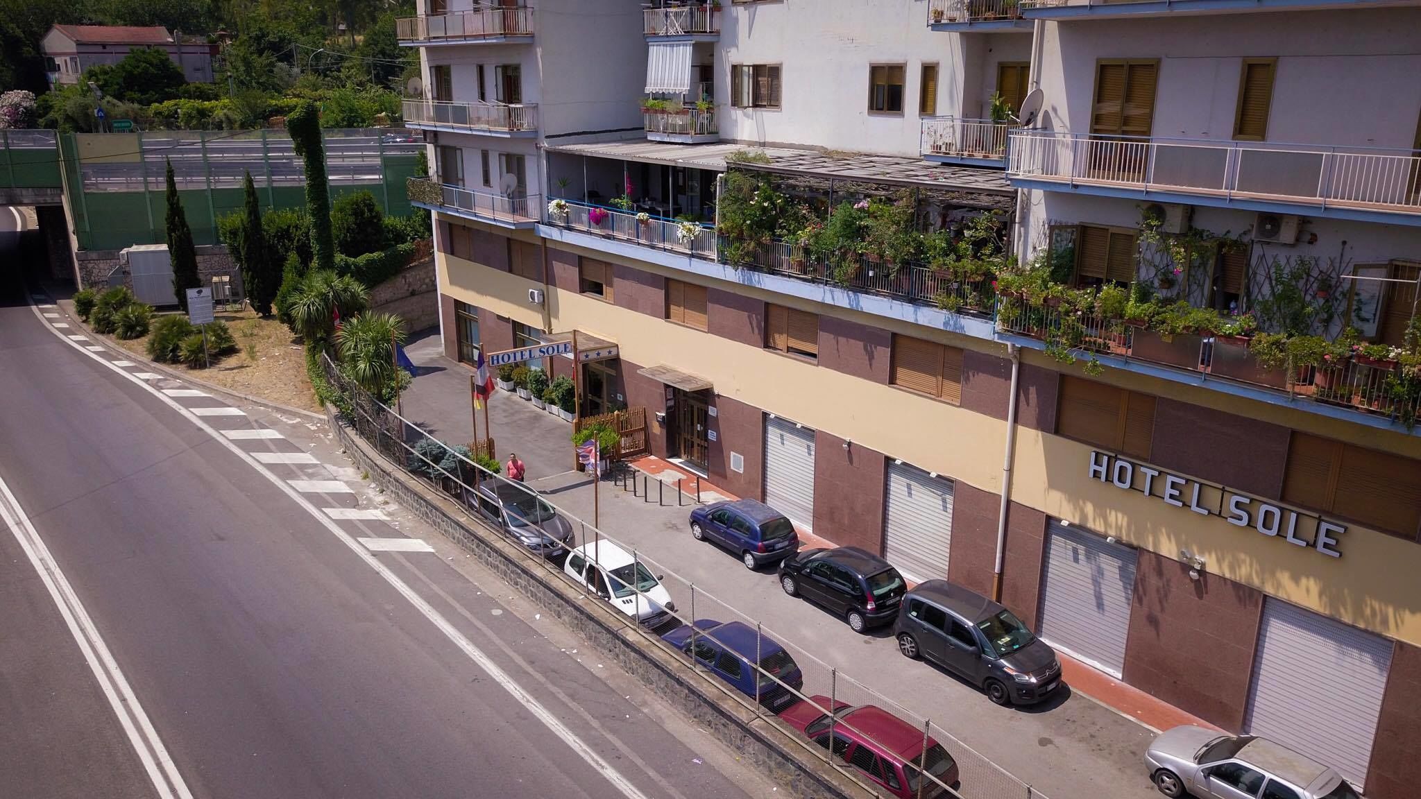 A hotel with multiple floors, balconies with plants, and a street view with parked cars.