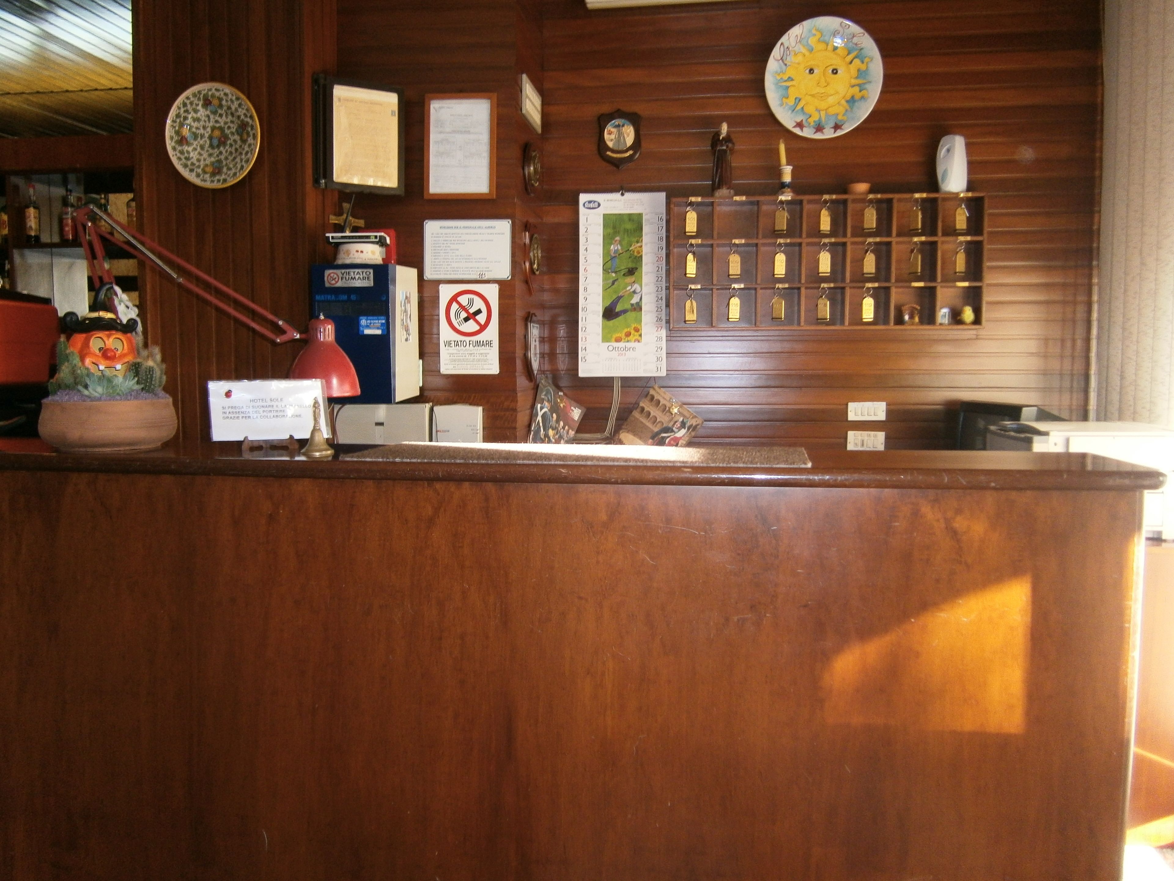 Un mostrador de recepción de madera con certificados, un calendario, un cartel de no fumar y un panel de tarjetas electrónicas en la pared.