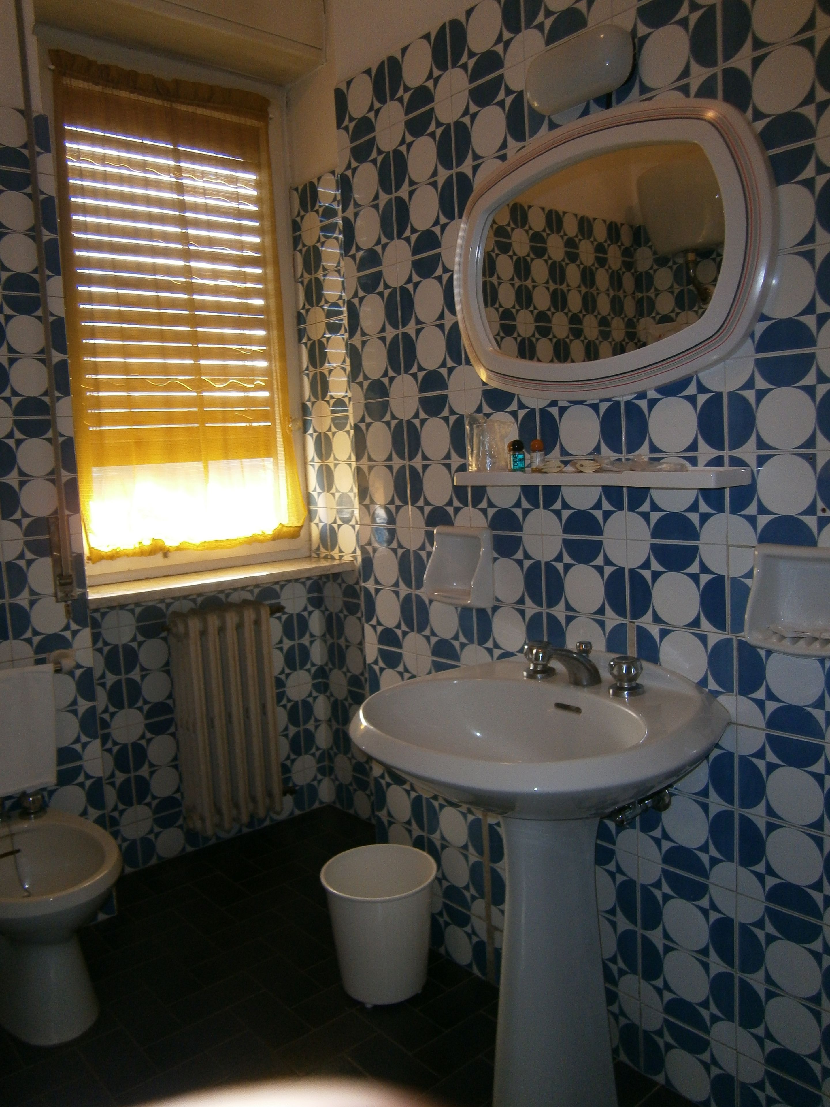 A bathroom with a patterned tile wall, a sink, a toilet, and a window with blinds.