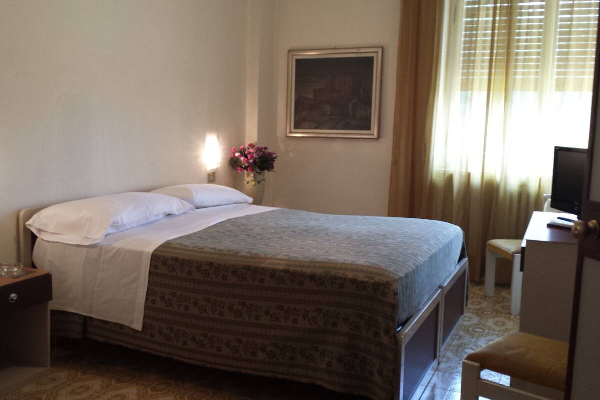 A hotel room with a bed, bedside table, lamp, and a painting on the wall.