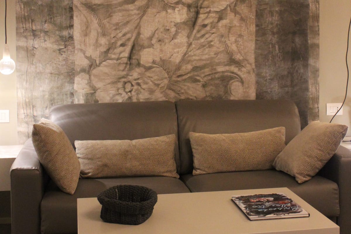 A living room with a large floral tapestry on the wall, a brown sofa with patterned cushions, and a white coffee table with a magazine and a basket.