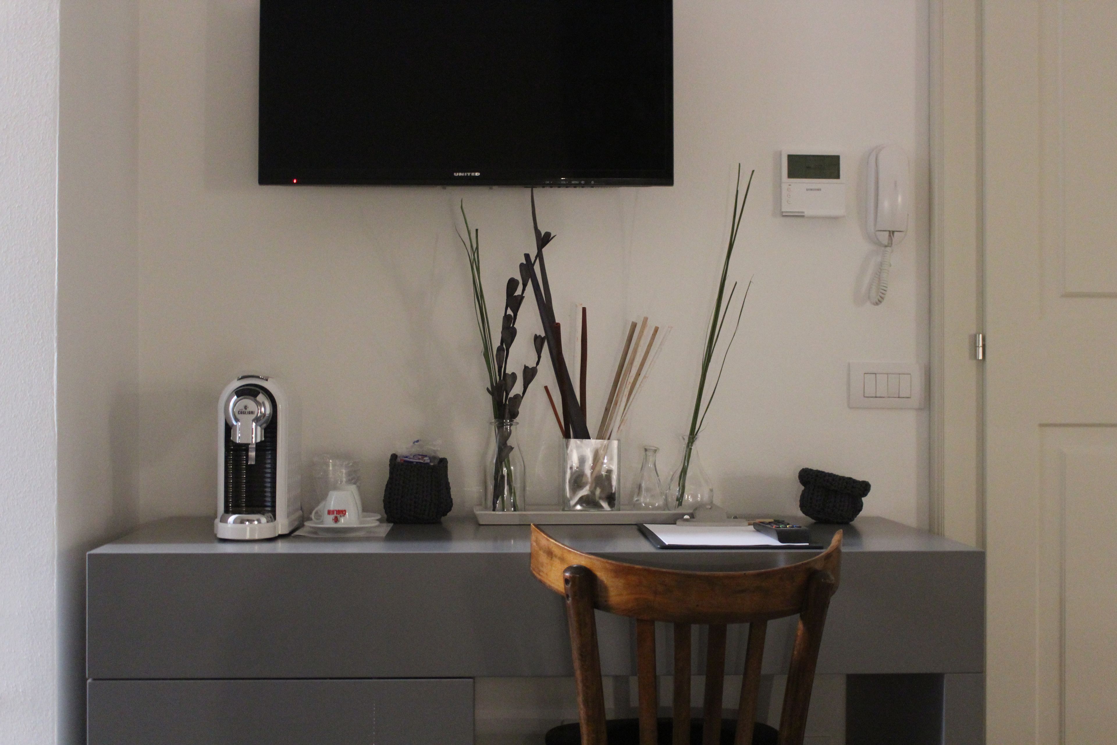 Een bureau met een flatscreen televisie, een telefoon en een koffiezetapparaat. Op het bureau staan decoratieve items, waaronder een vaas met takken en een glazen vaasje. Een stoel staat voor het bureau.