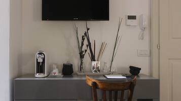 Een bureau met een flatscreen televisie, een telefoon en een koffiezetapparaat. Op het bureau staan decoratieve items, waaronder een vaas met takken en een glazen vaasje. Een stoel staat voor het bureau.