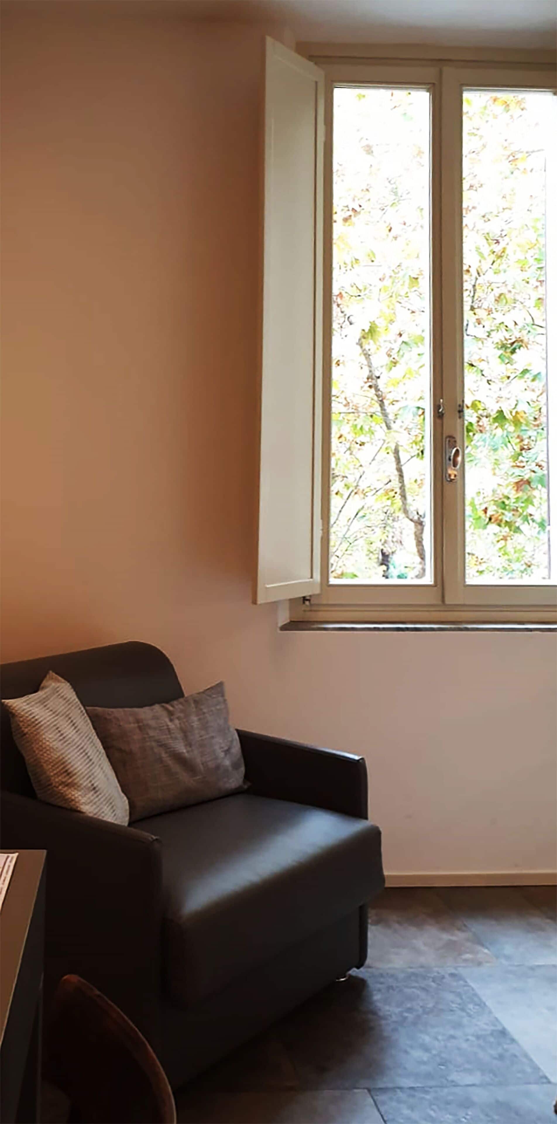 Una habitación con un sofá de color oscuro, un ventanal con vistas a árboles y un piso de baldosas.