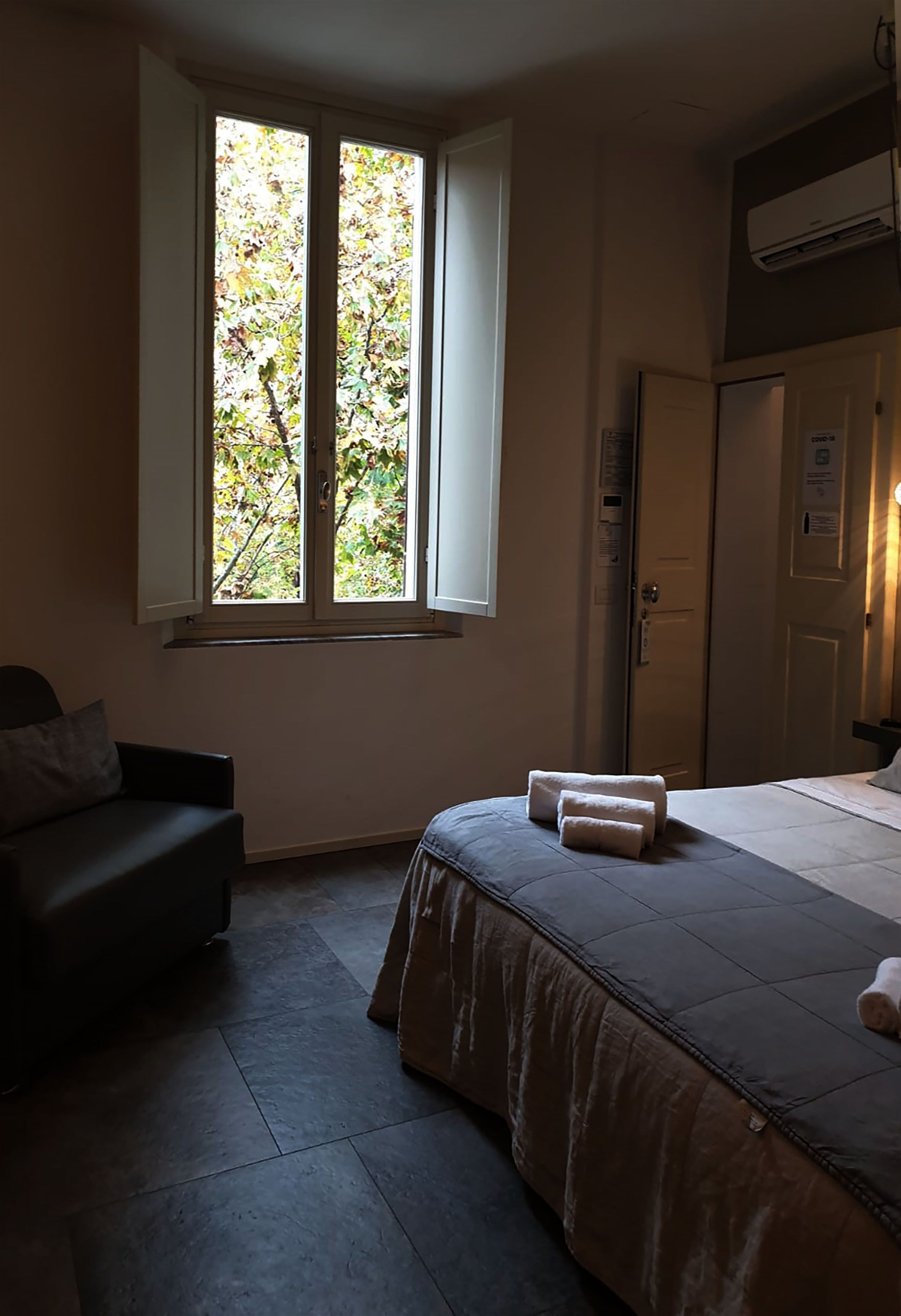 Quarto de hotel com cama, janela com vista para árvores, porta e pequena mesa com abajur.