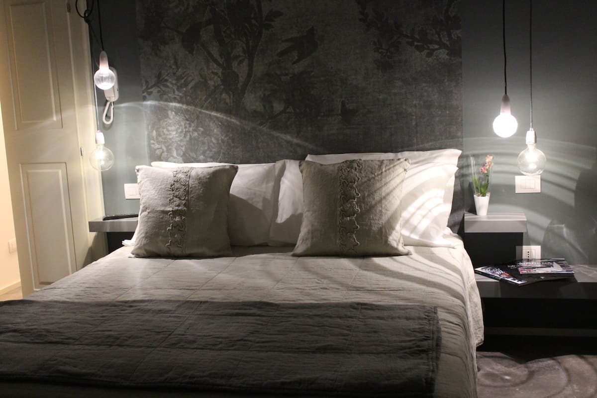 A neatly made bed with a white comforter and two decorative pillows, a bedside table with a magazine, and a wall with a floral pattern.