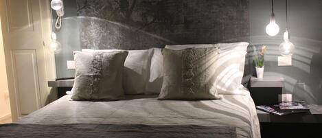 A neatly made bed with a white comforter and two decorative pillows, a bedside table with a magazine, and a wall with a floral pattern.
